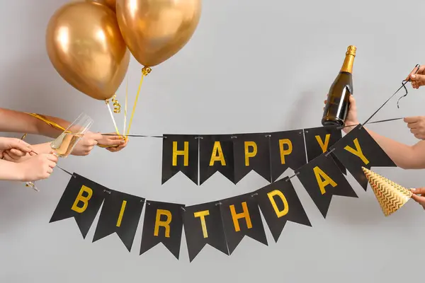 Female hands with birthday garland, bottle of champagne and balloons on light background
