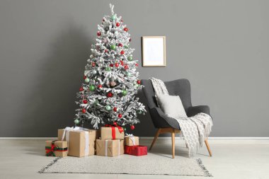 Gri duvara karşı hediyeler ve koltukla süslenmiş Noel ağacı.