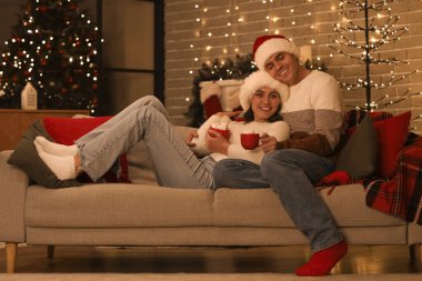 Genç çift Noel gecesi kanepede kremalı sıcak kakao içiyor.