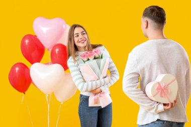 Hediye kutusu, çiçek buketi ve sarı arka planda kalp şeklinde balonları olan sevimli bir çift. Sevgililer Günü kutlaması
