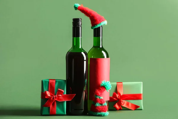 Bottles of wine with knitted hats and Christmas gift boxes on green background