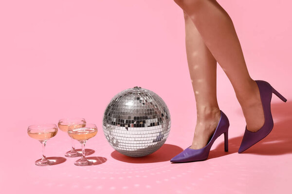 Female legs in high heel shoes with glasses of champagne and disco ball on pink background. New year party concept