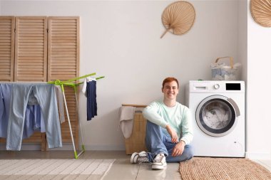Genç adam evde çamaşır makinesinin yanında oturuyor.