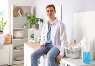 Cep telefonlu genç adam evde çamaşır makinesinin yanında oturuyor.