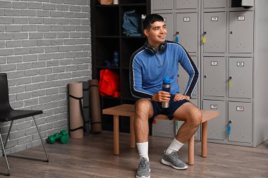 Soyunma odasındaki dolabın yanında oturan su şişeli genç adam.