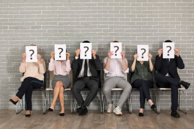 Adaylar, gri tuğla duvarın yanında soru işaretli kağıt kağıtlar taşıyorlar