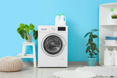 Çamaşır makinesi, raf ünitesi, ev bitkileri ve mavi duvarın yanındaki puf.