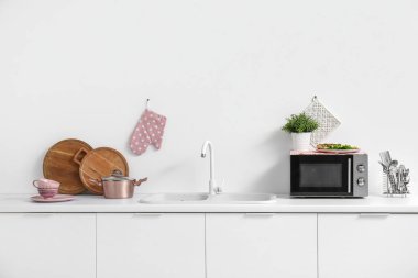 Beyaz tezgahlarda mikrodalga fırın, lavabo ve mutfak aletleri olan bir mutfak.