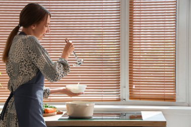 Genç bir kadın mutfakta tavuk çorbasını kaseye dolduruyor.