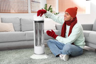 Genç adam elektrikli ısıtıcının yanında ısınıyor.