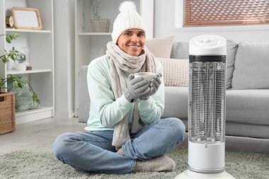 Evde elektrikli ısıtıcının yanında çay ısıtan genç bir adam.