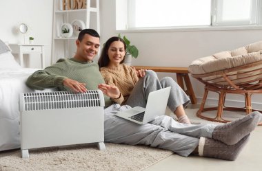 Yatak odasındaki radyatörün yanında laptoplu genç çift ısınıyor.