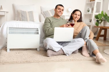 Yatak odasındaki radyatörün yanında laptoplu ve şaraplı genç çift ısınıyor.