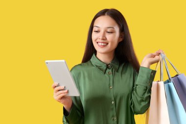 Tablet bilgisayarı ve sarı arka planda alışveriş torbaları olan genç Asyalı kadın.