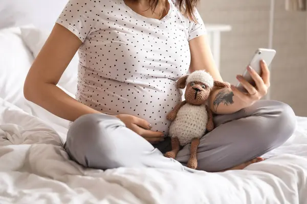 Young pregnant woman with toy using mobile phone in bedroom, closeup