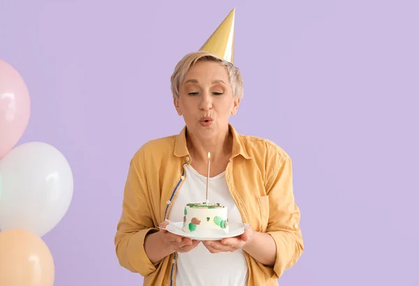 Mature woman with birthday cake making wish on lilac background