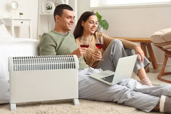 Young couple with wine and laptop warming near radiator in bedroom