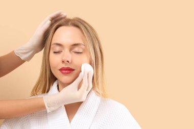 Pamuk yastıklı kozmetolog bej arka planda genç bir kadının yüzündeki makyajı temizliyor.