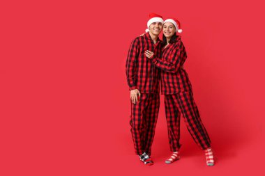 Mutlu genç çift Noel pijamaları ve Noel Baba şapkaları ile kırmızı arka planda sarılıyorlar.