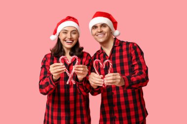 Mutlu genç çift, Noel pijamalarıyla pembe arka planda şeker kamışları yapıyorlar.