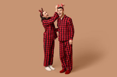 Mutlu genç çift Noel pijamalarıyla ve geyik boynuzlarıyla bej arka planda