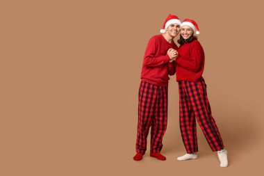 Mutlu genç çift, Noel pijamaları ve Noel Baba şapkaları ile bej arka planda sarılıyorlar.