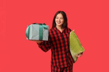 Kırmızı arka planda, yastıklı ve Noel hediye kutulu, kareli pijamalı güzel, mutlu bir Asyalı kadın.