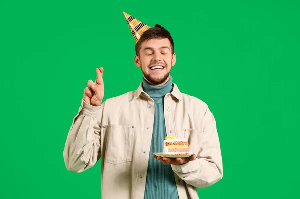 Young bearded man in party hat with piece of birthday cake making wish on green background