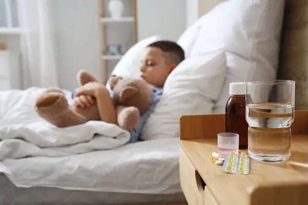 Pills with cough syrup and water on table of ill little boy in bedroom, closeup