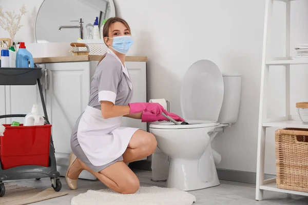 Female janitor with mask cleaning toilet bowl in bathroom