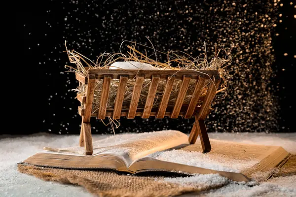 Bible, wooden manger with hay and dummy of baby on snow against black background. Concept of Christmas story
