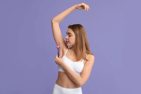 Beautiful young woman with razor shaving armpit on purple background