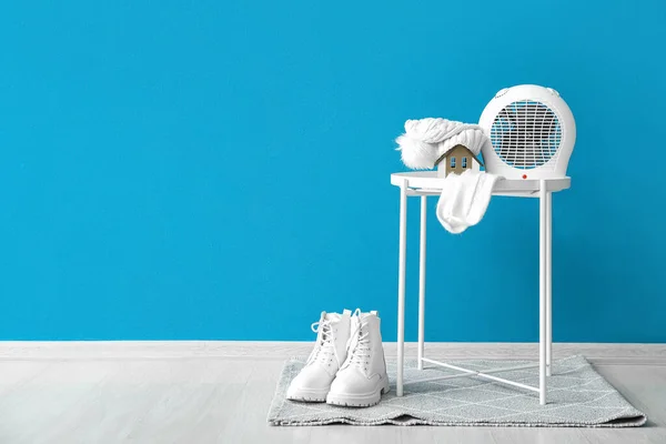 Electric fan heater with hat, socks on coffee table and boots near blue wall. Concept of heating season