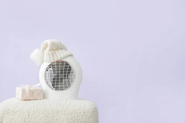 Electric fan heater with hat and gift box on pouf on lilac background. Concept of heating season