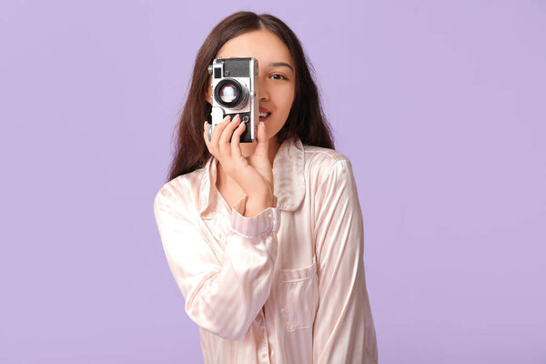 Красивая молодая счастливая азиатка в пижаме с камерой на фиолетовом фоне