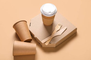 Paketlenmiş kağıt bardaklar, pizza kutusu ve bej arka planda çatal bıçak takımı.