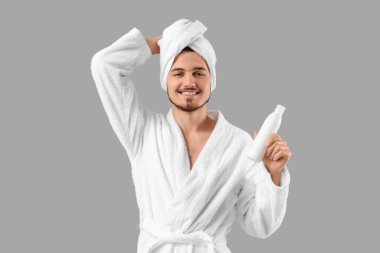 Gri arka planda şampuanı olan bornozlu yakışıklı bir genç.