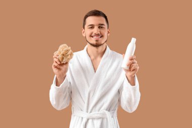 Banyo süngeri ve bej arka planda duş jeli ile bornozlu yakışıklı bir genç.