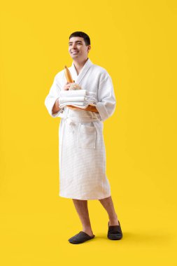 Sarı arka planda banyo aksesuarları olan bornozlu yakışıklı bir genç.