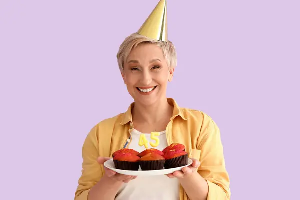 Mature woman with birthday muffins on lilac background