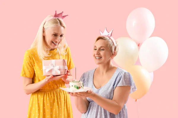Mature women with birthday cake and gift on pink background