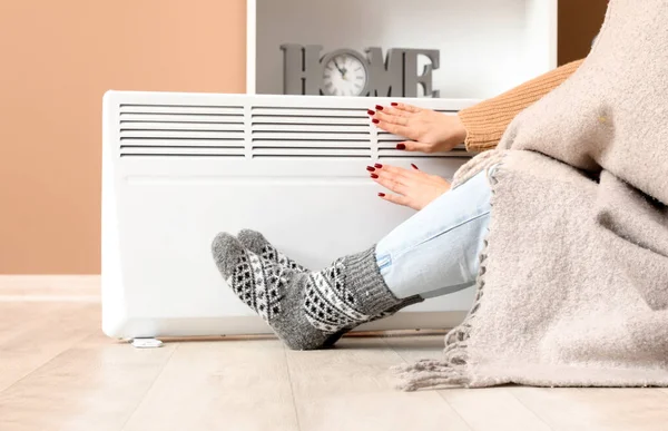 Young woman in socks with warm plaid near electric heater at home. Concept of heating season