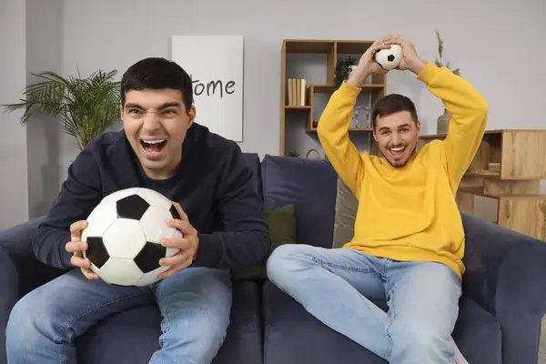 Handsome young brothers sitting on sofa with football balls in living room