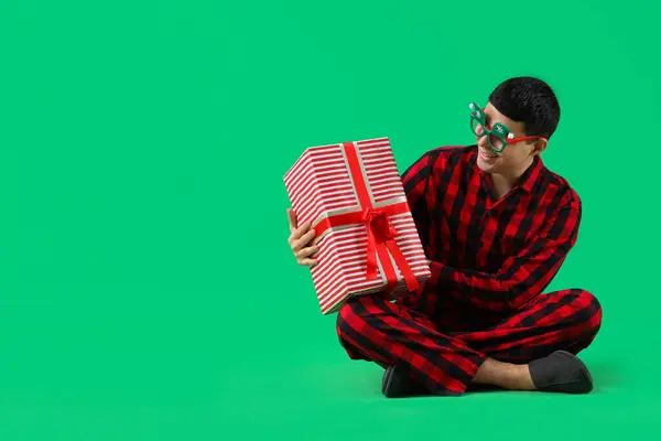 Young man in pajamas with Christmas gift box on green background
