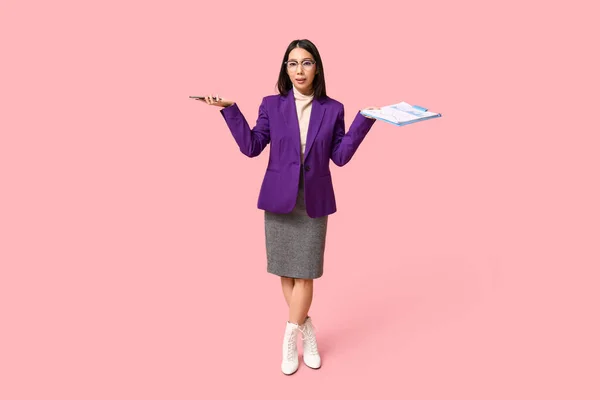 Portrait of young Asian businesswoman with mobile phone and clipboard shrugging on pink background