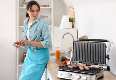 Mutfaktaki modern elektrikli ızgarada lezzetli sosisler pişirirken tablet bilgisayar kullanan güzel genç bir kadın.