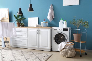 Çamaşır odasının içinde çamaşır makinesi, beyaz tezgah ve kuru askılı kıyafetler var.