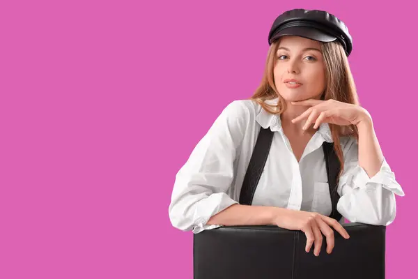 Stylish young woman with suspenders sitting in chair on purple background