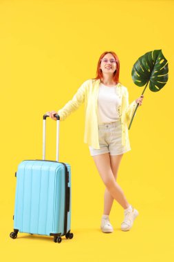 Sarı arka planda bavulu ve tropikal yaprağı olan kadın turist.