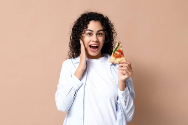 Güzel, genç, şok olmuş, bej arka planda bir dilim lezzetli pizzayla Afro-Amerikan kadın.